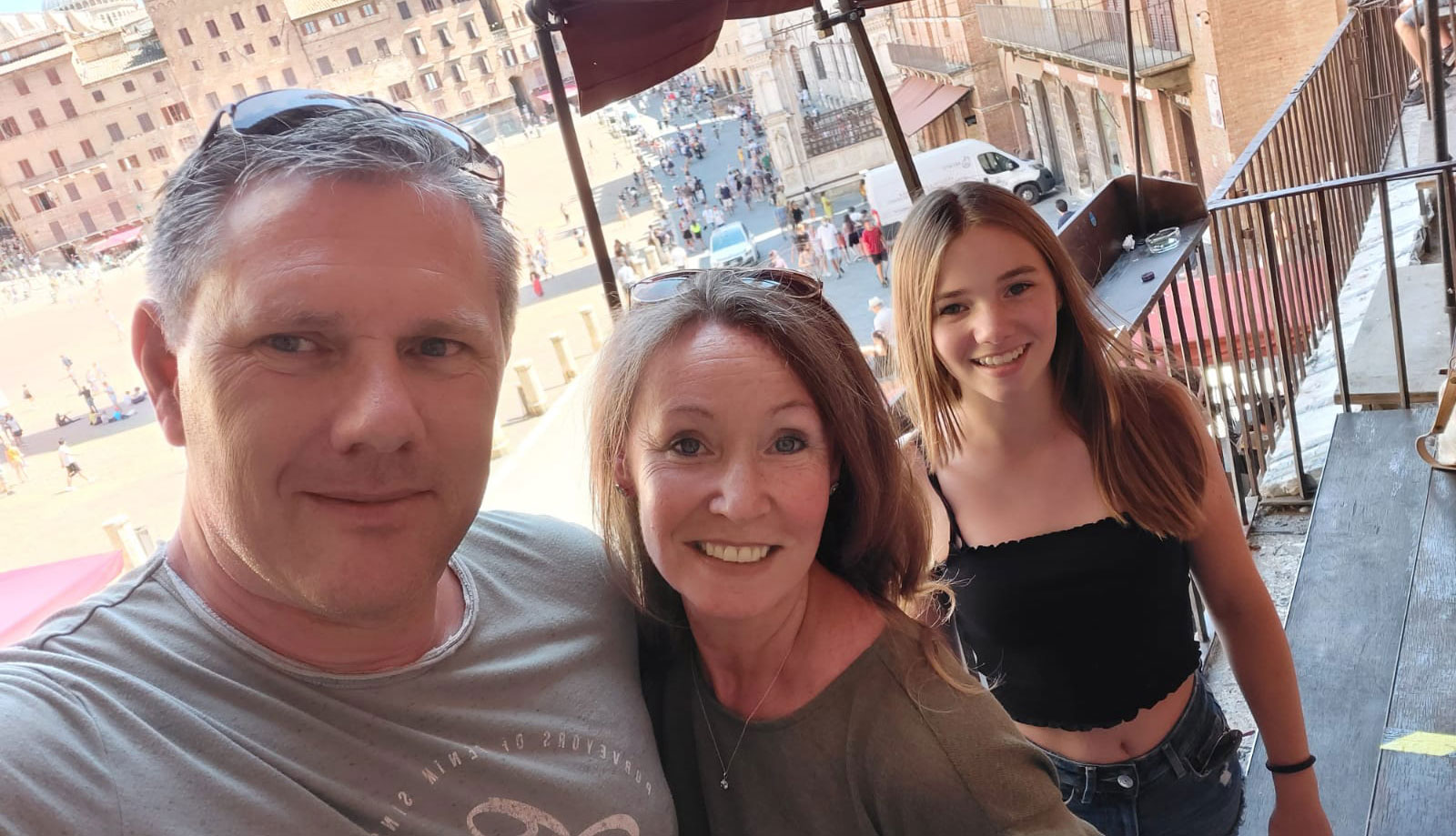 Ein Familienfoto aus dem Sommerurlaub: Im Vordergrund ein Mann, der das Selfie macht, daneben seine Frau und die Tochter. Im Hintergrund sind ein großer Platz und eine mediterrane Stadt zu erkennen.