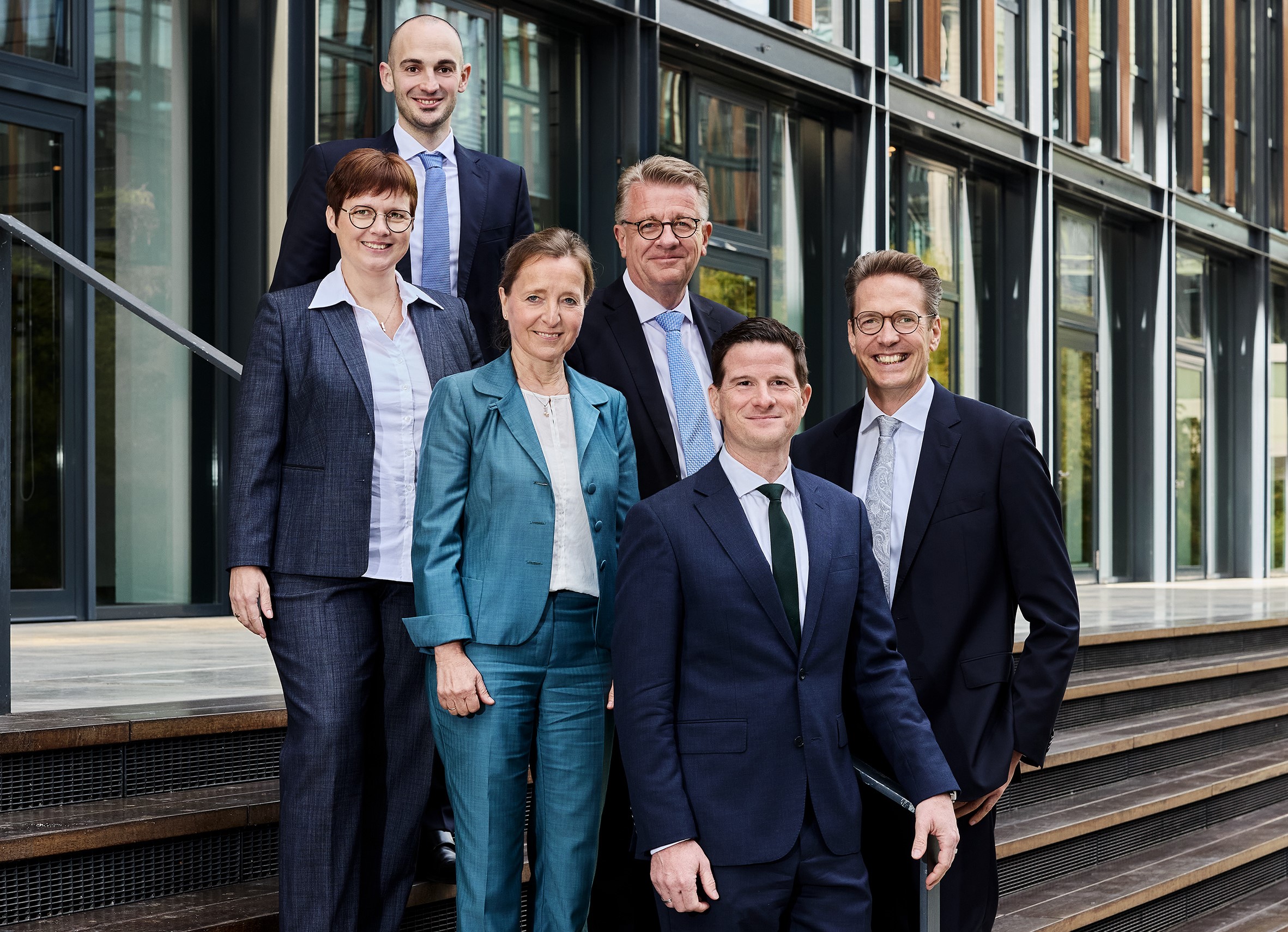 Eine Gruppe von vier Männern und zwei Frauen in Business-Kleidung auf einer Treppe vor einem Bürogebäude. Sie stehen versetzt auf den Stufen und lächeln in die Kamera.