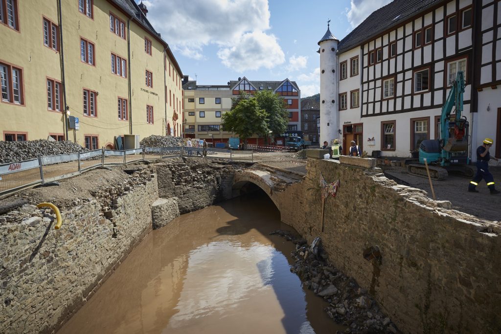 Eine zerstörte Straße über eine gemauerte Brücke in Bad Münstereifel - aufgenommen zwei Wochen nach der Flutkatastrophe 2021. Daneben alte Häuser, teils mit Fachwerk, in der historischen Innenstadt. Ein Bagger steht am Rand und Hilfskräfte sind unterwegs.