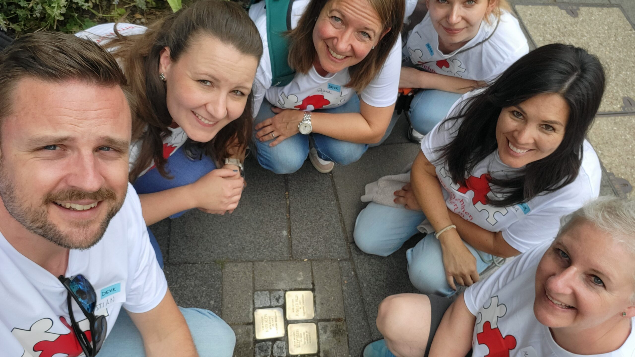 Gruppenbild mit fünf Frauen und einem Mann. Alle tragen dieselben weißen T-Shirts zur Aktion "Unternehmen engagiert in Köln" und knien am Boden, wo sie sich im Halbkreis um glänzende Stolpersteine versammelt haben. Sie blicken nach oben Richtung Kamera.
