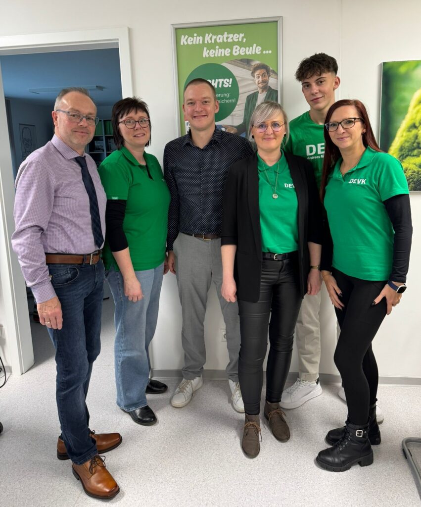 Gruppenbild mit drei Männern und drei Frauen in einer DEVK-Geschäftsstelle. Vier Personen tragen grüne Poloshirts mit DEVK-Logo, die anderen Hemden. Im Hintergrund ist eine Kfz-Werbung der DEVK als Plakat an der Wand zu sehen.