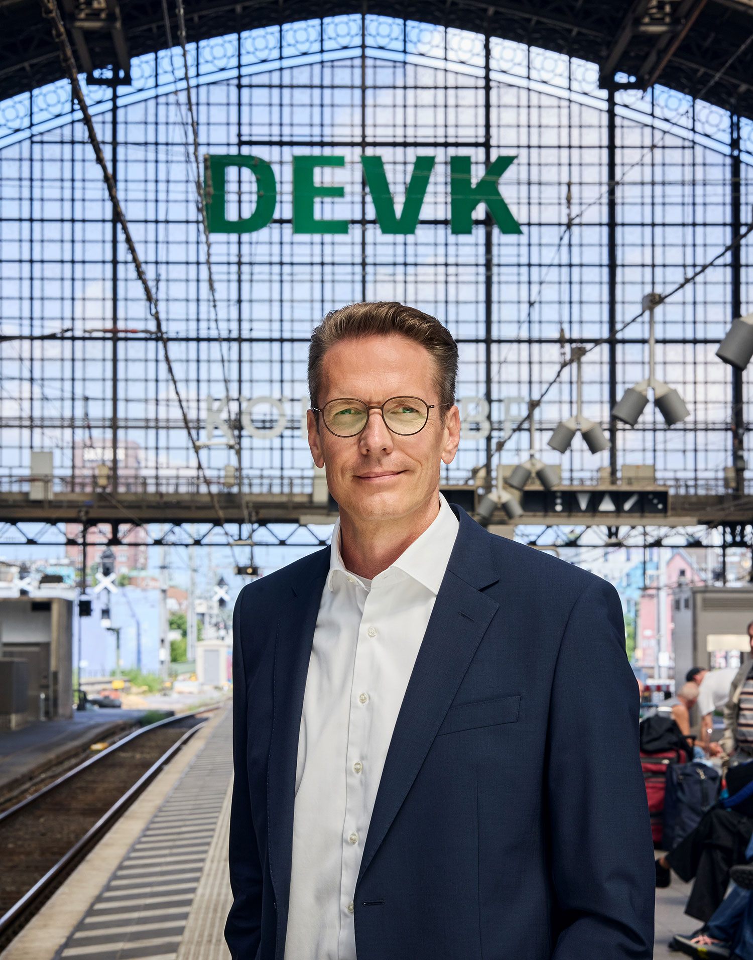 Ein Mann mit Brille in dunkelblauem Anzug und weißem Hemd steht an einem Gleis im Kölner Hauptbahnhof und lächelt in die Kamera. Hinter ihm verlaufen Gleise, über ihm sind die Buchstaben DEVK als Leuchtwerbeanlage unter dem gewölbten Bahnhofsdach zu sehen.