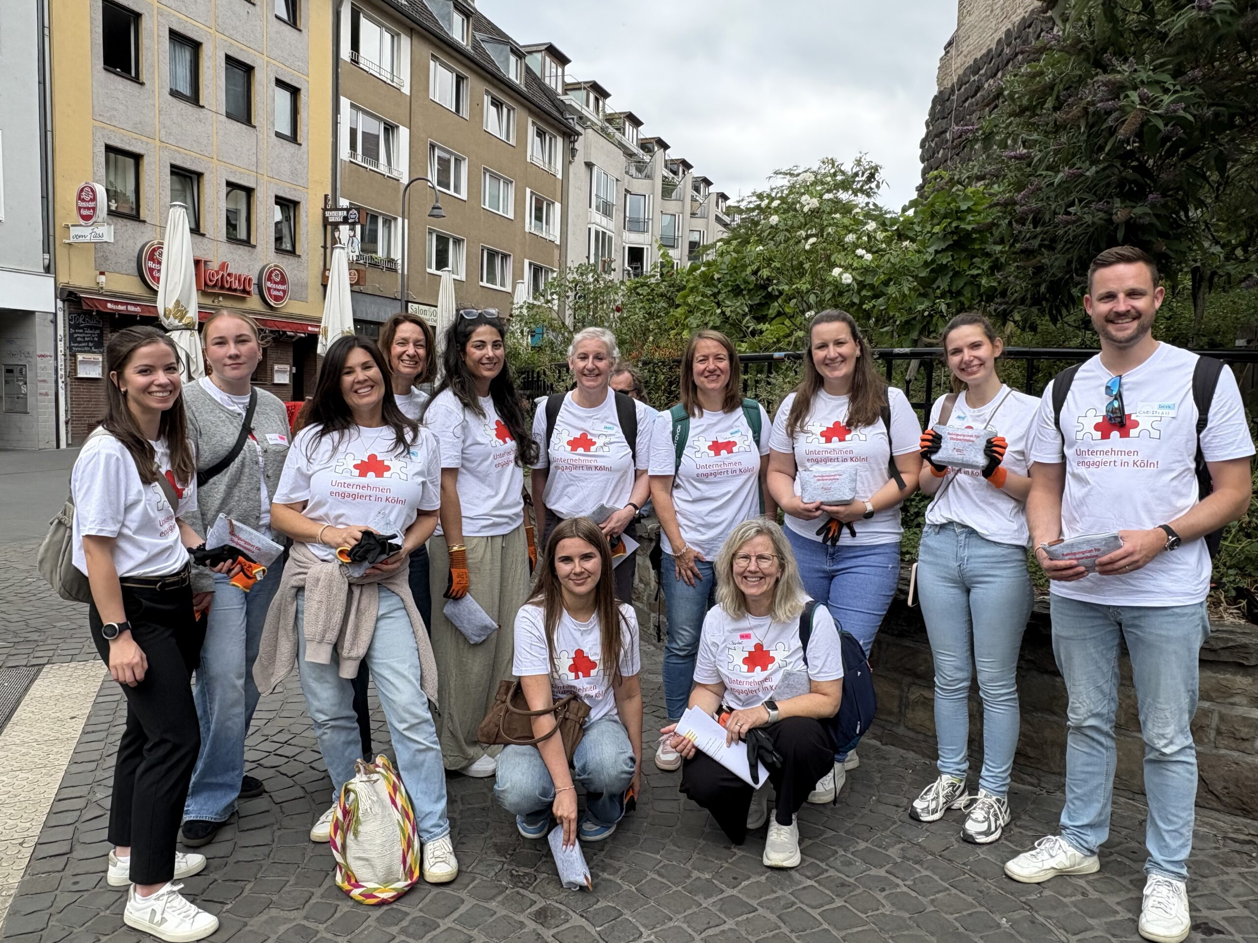 Gruppenbild mit elf Frauen und einem Mann in der Innenstadt von Köln. Alle tragen die gleichen weißen T-Shirts mit drei verbundenen Puzzlestücken und der Aufschrift "Unternehmen engagiert in Köln".