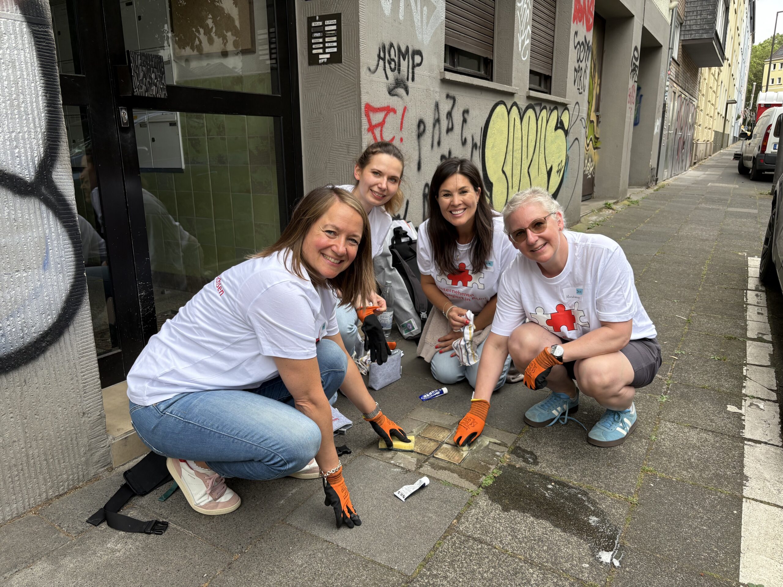 Vier Frauen hocken auf dem Boden vor einem mit Graffiti besprühten Haus in Köln und putzen Stolpersteine. Sie tragen dabei orangefarbene Handschuhe.