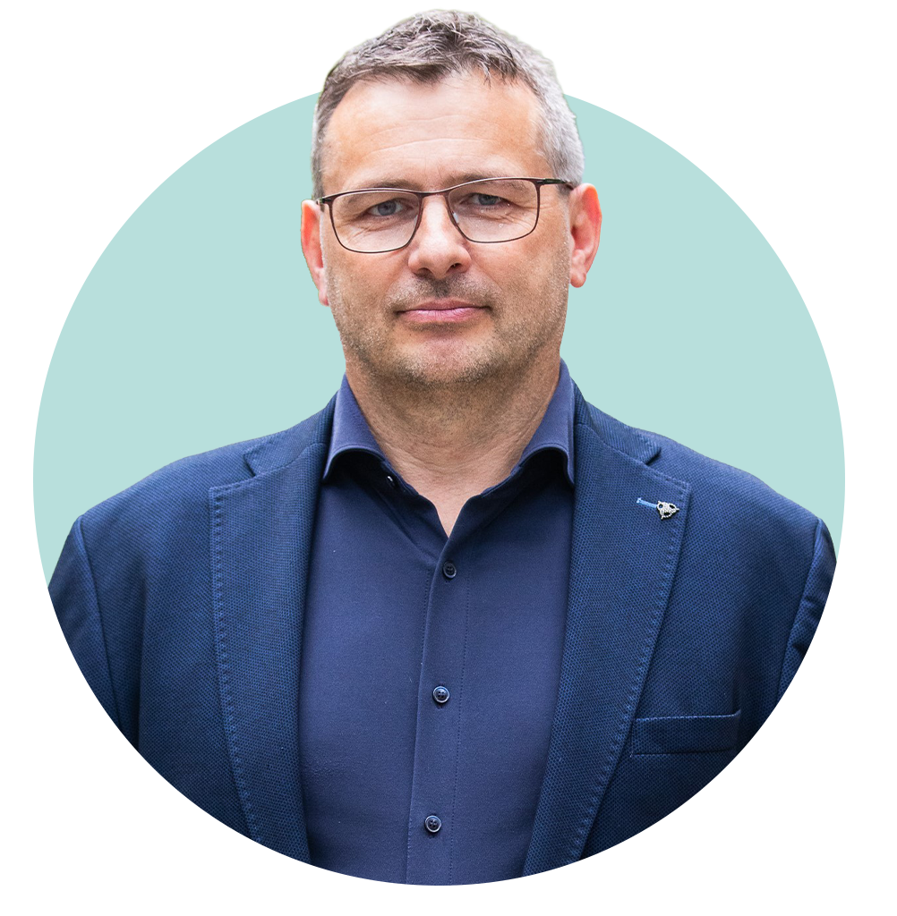 Ein Mann mit grauen Haaren in dunkelblauem Anzug und dunkelblauem Hemd lächelt in die Kamera. Im Hintergrund sind grüne Bäume zu sehen.