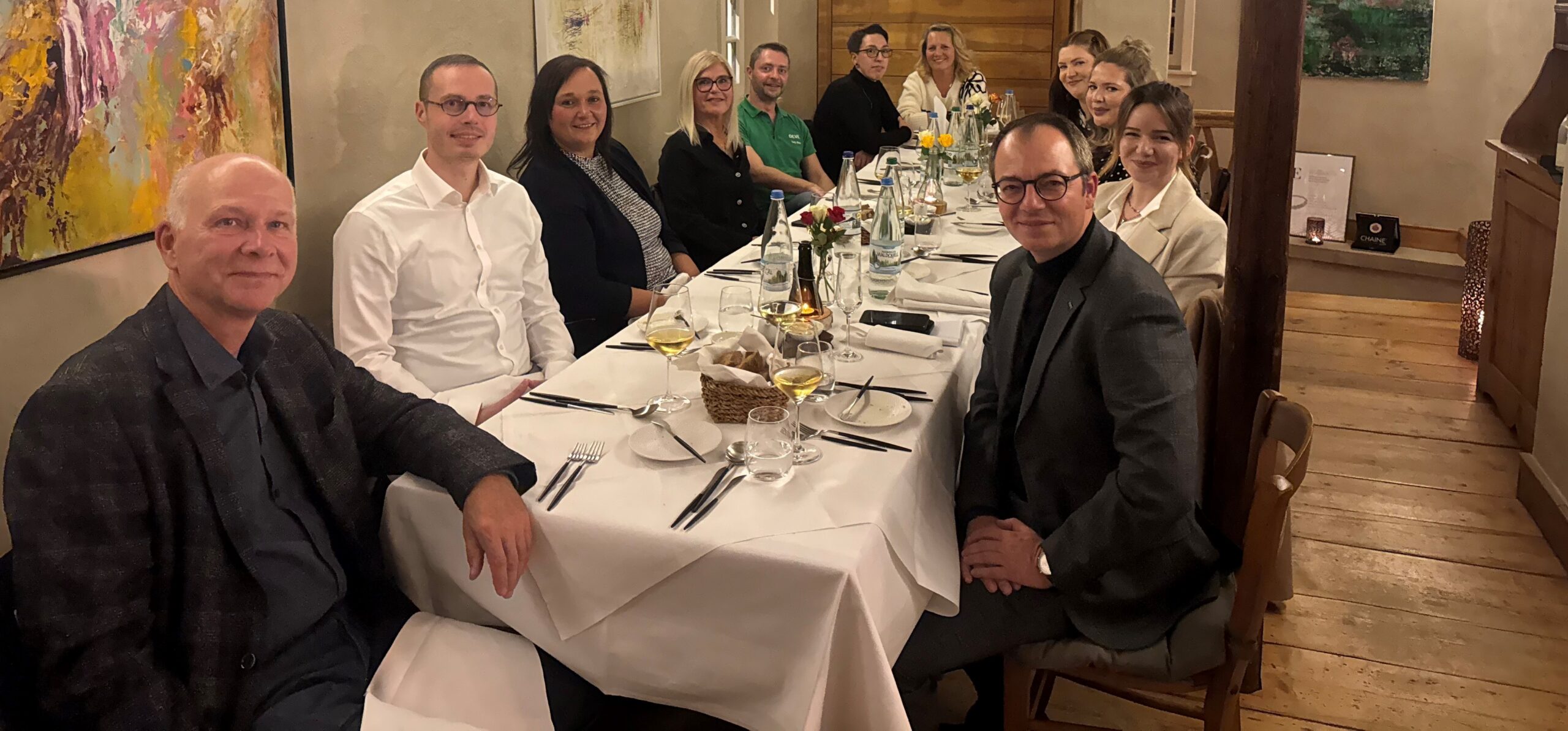 An einer langen Tafel mit weißer Tischdecke sitzen vier Männer und sieben Frauen in Businesskleidung in einem Restaurant am gedeckten Tisch und lächeln in die Kamera.
