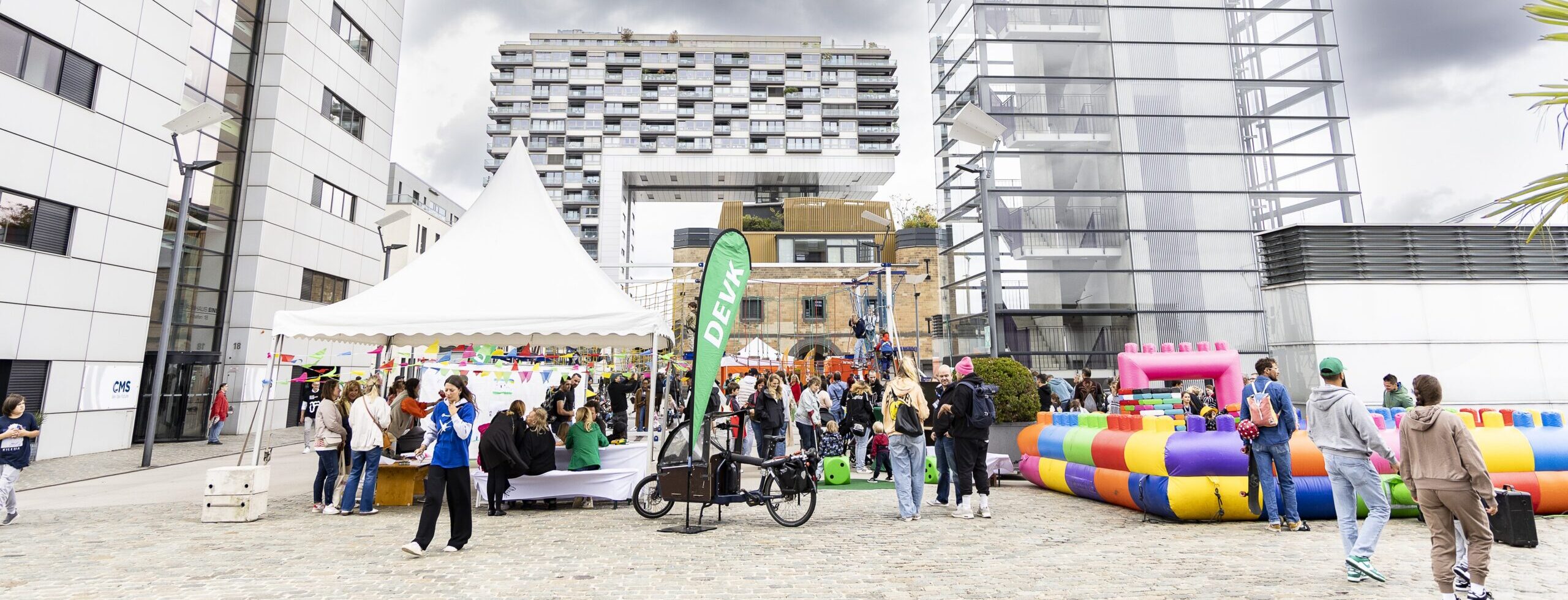 Auf der Fläche vor den Kölner Kranhäusern ist ein weißes Zelt mit einem Infostand aufgebaut, eine Beachflag mit DEVK-Logo lädt die Familien zum Weltkindertag ein und rechts im Bild befindet sich ein Parcours für Kinder mit einem bunten Pufferrand in Klemmbaustein-Optik.